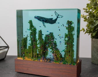 May include: A rectangular resin art piece featuring a whale swimming above a detailed underwater scene. The scene includes colorful coral and fish, set on a wooden base. The resin is clear, creating a 3D effect.