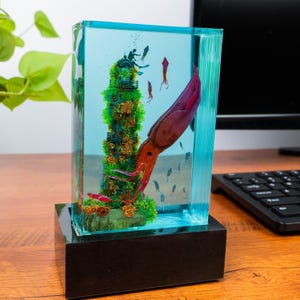 May include: A rectangular resin sculpture featuring an underwater scene with a giant squid, divers, and coral. The sculpture is encased in a clear, turquoise-colored block, mounted on a black base. The squid is a deep red and purple color.
