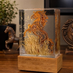 May include: A rectangular clear resin sculpture featuring a rearing horse in shades of gold and brown, set against a backdrop of dried grass. The sculpture is mounted on a wooden base, creating a striking decorative piece.