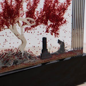 May include: A decorative diorama featuring a Japanese scene with a white-trunked tree with red leaves, a black tombstone, and a silhouetted figure. The scene is encased in a clear acrylic box with a black base.