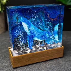 May include: A blue whale sculpture encased in clear resin, set on a wooden base. The whale is painted in shades of blue, with bubbles and a textured background. The artwork is a decorative piece.