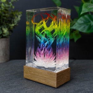 以下が含まれることがあります： 虹色のデザインが施された、長方形の透明樹脂製アート作品。木製のベースと、鮮やかでマルチカラーのインテリアデザインが特徴です。色は赤から紫へと変化し、印象的な視覚効果を生み出しています。装飾品です。