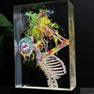 May include: A clear acrylic sculpture featuring a skeletal figure with colorful paint splatter. The skeleton is depicted in a dynamic pose, with vibrant hues of yellow, green, blue, and orange. The sculpture is set against a black background.