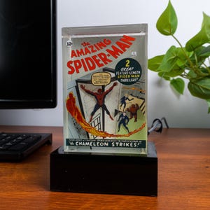May include: A clear acrylic block featuring a vintage comic book cover of The Amazing Spider-Man. The cover art depicts Spider-Man with other characters and the text "The Chameleon Strikes!" on a black base.