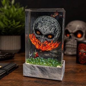 May include: A rectangular resin art piece featuring a detailed, ominous moon with fiery accents and glowing eyes. The artwork includes a miniature scene with a figure and flowers, set on a stone-like base. The background is a starry night.