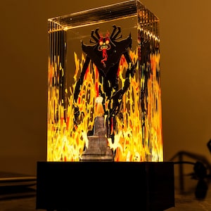 Peut inclure: Un présentoir rectangulaire éclairé avec une silhouette noire d'une figure de démon cornu avec un visage rouge et une petite figurine en robe blanche. Le fond est un motif flamboyant orange et jaune, le tout enfermé dans un bloc acrylique transparent.