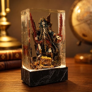 May include: A detailed pirate figurine inside a clear, rectangular display case. The pirate skeleton wears a tricorn hat and tattered coat, holding two red swords. A treasure chest sits at the base. The display case rests on a black marble-like base.