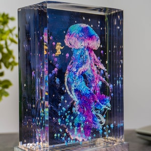 May include: A rectangular resin art piece featuring a vibrant, colorful jellyfish design. The artwork includes an astronaut and smaller jellyfish against a dark blue, starry background. The jellyfish is primarily blue, purple, and pink.
