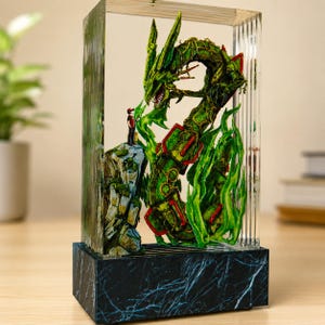 May include: A detailed resin sculpture of a green dragon with red accents, encased in a clear rectangular block. A small figure stands on a rocky outcropping, facing the dragon. The base is a dark, marbled pattern.