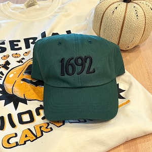May include: A dark green baseball cap with a black embroidered number "1692" on the front. The cap is lying on a white t-shirt with a pumpkin graphic and the text "SERIOUSLY, I LOVE CARVING".