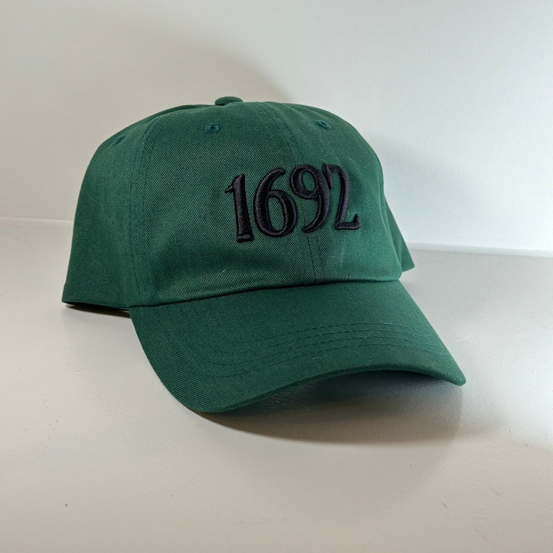 May include: A dark green baseball cap with black embroidered text that reads "1692".