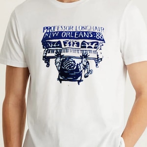 May include: White t-shirt featuring a blue graphic print of a person playing a piano. The text "Professor Longhair New Orleans: 88" is printed above the piano.