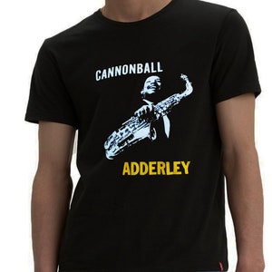 Może przedstawiać: Czarny T-shirt z niebiesko-białym nadrukiem przedstawiającym mężczyznę grającego na saksofonie. Na koszulce nadrukowany jest napis "Cannonball Adderley" w kolorze żółtym.