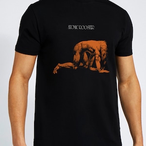 Könnte beinhalten: Schwarzes T-Shirt mit dem Text "ATOMIC ROOSTER" über einer orangefarbenen Illustration einer knienden Figur. Das kurzärmelige Shirt ist aus einem weichen Material gefertigt.