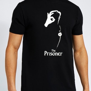 Könnte beinhalten: Schwarzes T-Shirt mit einer weißen Grafik einer Hand, die die "Nummer sechs" mit einem Kreis um Zeigefinger und Daumen zeigt. Der Text "The Prisoner" befindet sich unter der Grafik.