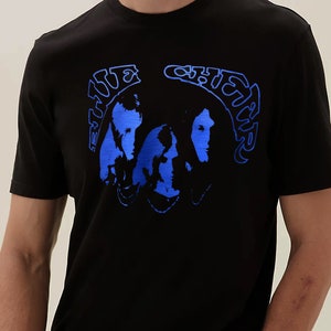 Può includere: Maglietta nera con una grafica blu dei volti dei membri della band The Beatles e il testo "The Beatles" in un font blu curvo.