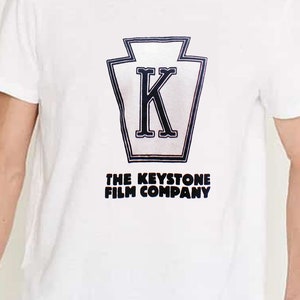 May include: White t-shirt featuring a black and white graphic of a Keystone Film Company logo. The logo depicts a stylised letter "K" within a shield shape.