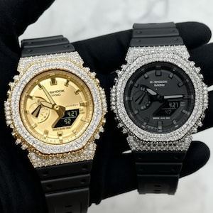 May include: Two G-Shock Casio watches with black resin bands and octagonal bezels encrusted with clear stones. One watch features a gold-toned dial, while the other has a black dial. Both displays show digital time and date information clearly.