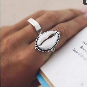 May include: Silver ring with a white seashell design. The ring is on a finger wearing a silver band.