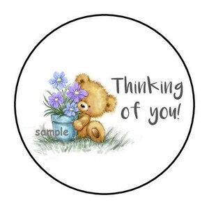 May include: A round sticker with a cartoon teddy bear holding a blue flower pot with purple and blue flowers. The text "Thinking of you!" is written in black, with the word "sample" at the bottom.