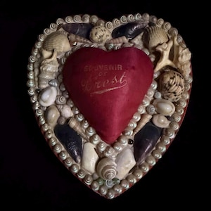 May include: Heart-shaped box decorated with seashells and a central red heart. The heart has gold text that reads "Souvenir of Crest". The box is trimmed with small, iridescent beads and various seashells.