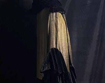 May include: A vintage garment displayed on a mannequin. The long skirt is made of a textured gold fabric. A black velvet cape is draped over the shoulders, with a matching velvet piece below. The background is a dark, solid colour.