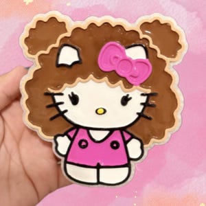 May include: A flat, shaped character charm featuring a white cat face with black whiskers, a yellow nose, and a bright pink bow. The character has brown, curly hair and is wearing a pink shirt with two black circular details on the front.