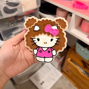 May include: A flat, decorative acrylic charm featuring a cartoon character with white skin, black whiskers, and a yellow nose. The character has brown, curly hair styled in two buns, wears a bright pink bow, and a matching pink shirt with two dots.
