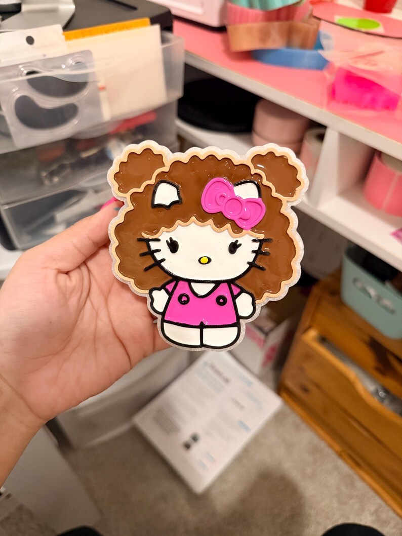 May include: A flat, decorative acrylic charm featuring a cartoon character with white skin, black whiskers, and a yellow nose. The character has brown, curly hair styled in two buns, wears a bright pink bow, and a matching pink shirt with two dots.