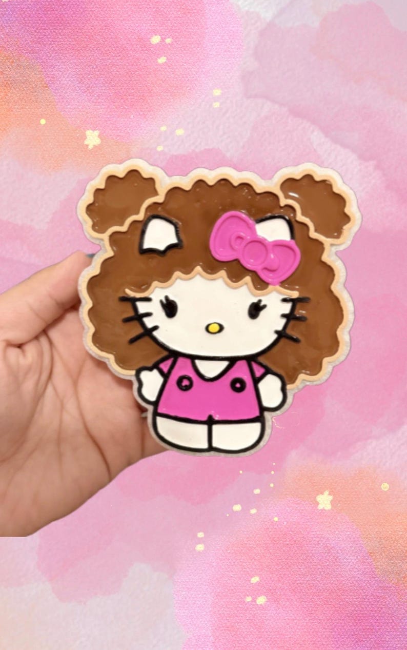 May include: A flat, shaped character charm featuring a white cat face with black whiskers, a yellow nose, and a bright pink bow. The character has brown, curly hair and is wearing a pink shirt with two black circular details on the front.