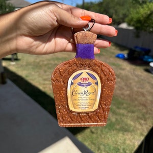 May include: A brown and gold Crown Royal bottle charm with a purple label. The charm is made of resin and has a textured surface. The label features the Crown Royal logo and the text "Fine de Luxe - Blended Canadian Whisky".