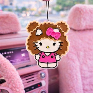 May include: A hanging car air freshener featuring a cartoon character with white skin, black eyes, and whiskers, wearing a bright pink shirt and a matching pink bow. The character has voluminous brown curly hair and is suspended by a black string.