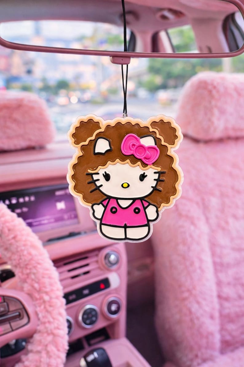 May include: A hanging car air freshener featuring a cartoon character with white skin, black eyes, and whiskers, wearing a bright pink shirt and a matching pink bow. The character has voluminous brown curly hair and is suspended by a black string.