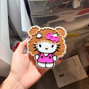 May include: A Hello Kitty character charm featuring a white face with black whiskers, eyes, and nose. The character has brown curly hair, a bright pink bow, and wears a pink shirt with two black buttons. The charm has a white border outline.