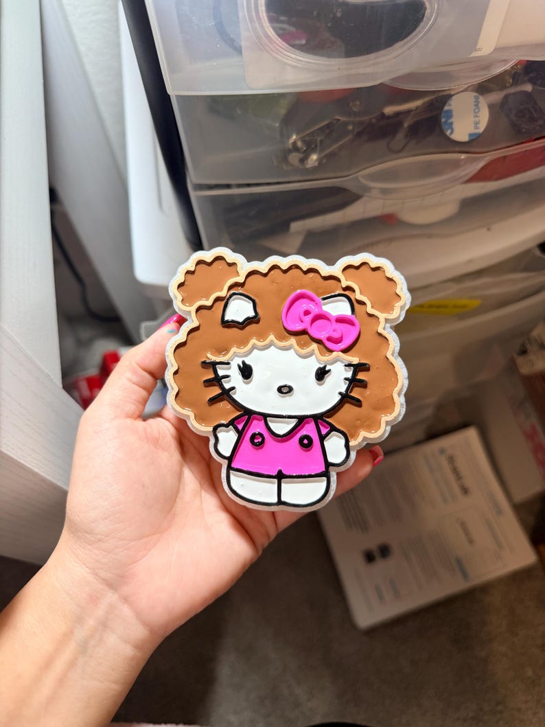 May include: A Hello Kitty character charm featuring a white face with black whiskers, eyes, and nose. The character has brown curly hair, a bright pink bow, and wears a pink shirt with two black buttons. The charm has a white border outline.