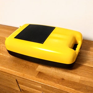 May include: A yellow plastic container with a black lid and a black handle. The container has a textured surface and a black vent on the top.