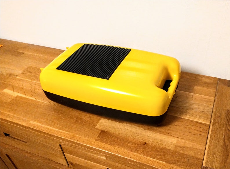 May include: A yellow plastic container with a black lid and a black handle. The container has a textured surface and a black vent on the top.