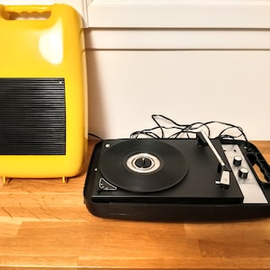May include: A vintage yellow record player with a black speaker and a black turntable with a silver tone arm. The record player is sitting on a wooden surface.