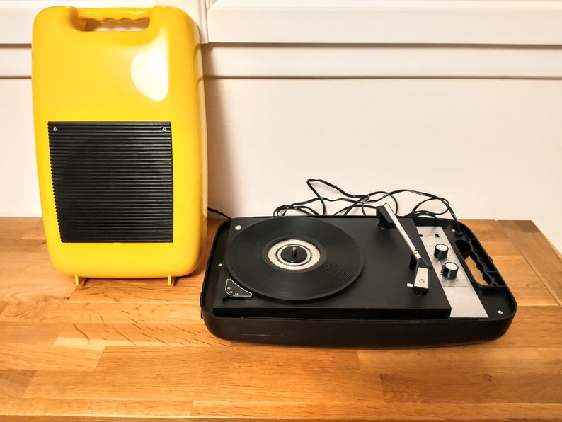May include: A vintage yellow record player with a black speaker and a black turntable with a silver tone arm. The record player is sitting on a wooden surface.