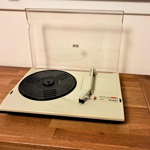 Puede incluir: Un tocadiscos vintage blanco con una tapa de plástico transparente. El plato giratorio es negro y tiene un brazo plateado.