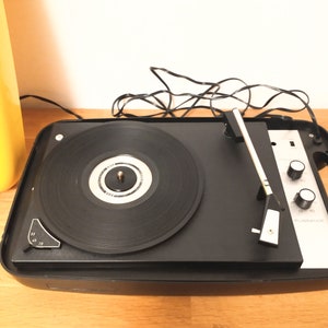 May include: A black vintage record player with a silver faceplate and a black tonearm. The record player has two knobs on the front and a handle on the side. The record player is sitting on a wooden surface.