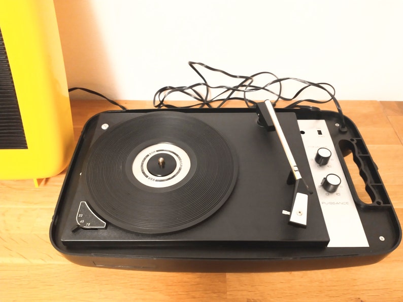 May include: A black vintage record player with a silver faceplate and a black tonearm. The record player has two knobs on the front and a handle on the side. The record player is sitting on a wooden surface.