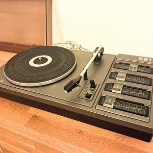 May include: A vintage Philips 381 turntable in a grey finish. The turntable has a black platter, tone arm, and control panel with sliders. The unit is set on a wooden surface.