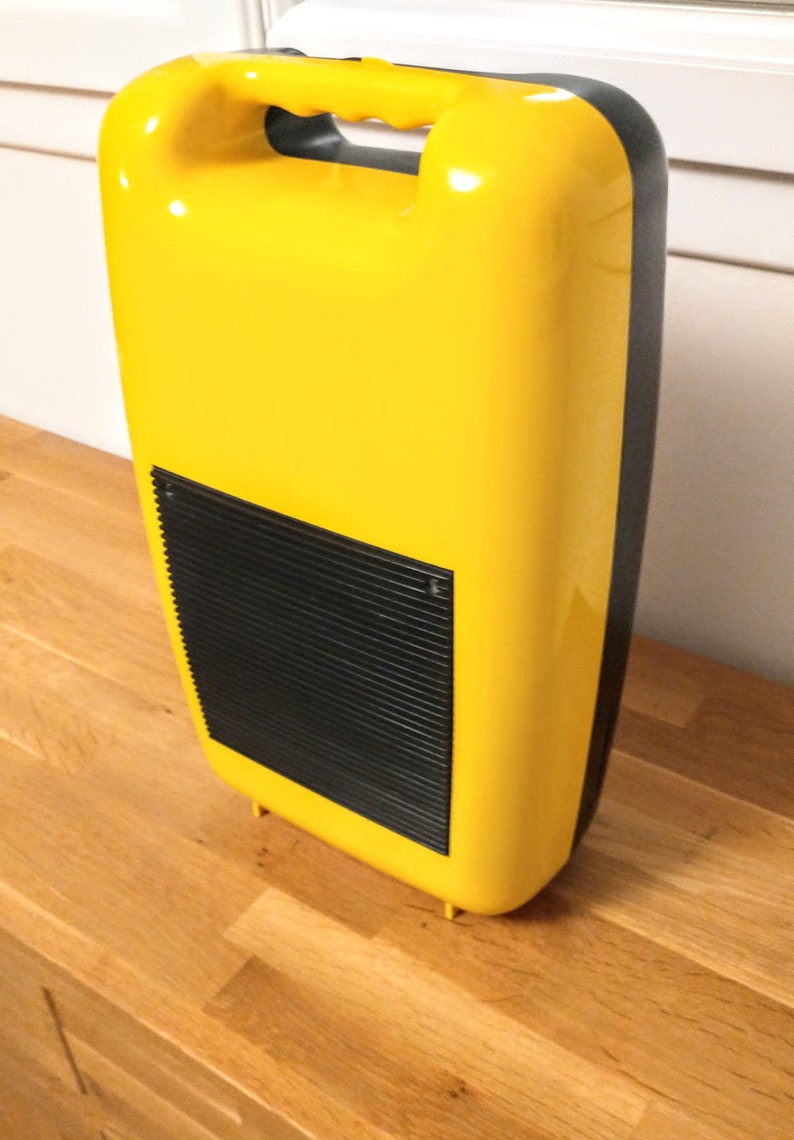 May include: A yellow plastic case with a black handle and a black grill on the front. The case has a rounded shape and a smooth surface.