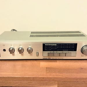 Puede incluir: Receptor estéreo vintage plateado con múltiples perillas, botones y un panel de visualización. La parte superior tiene ranuras de ventilación. El receptor está sobre una superficie de madera. La marca y el número de modelo son visibles.