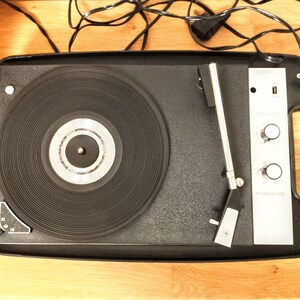 May include: A black portable record player with a silver control panel and a silver tonearm. The record player has a built-in speaker and a carrying handle.