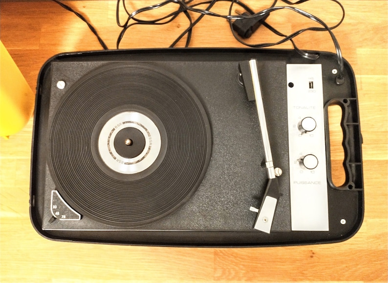 May include: A black portable record player with a silver control panel and a silver tonearm. The record player has a built-in speaker and a carrying handle.
