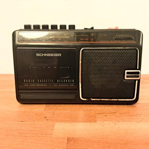 Puede incluir: Grabadora de casete de radio Schneider negra con altavoz, dial de sintonización y botones de control. El texto "Schneider" y "Radio Cassette Recorder" son visibles. El dispositivo está sobre una superficie de madera.