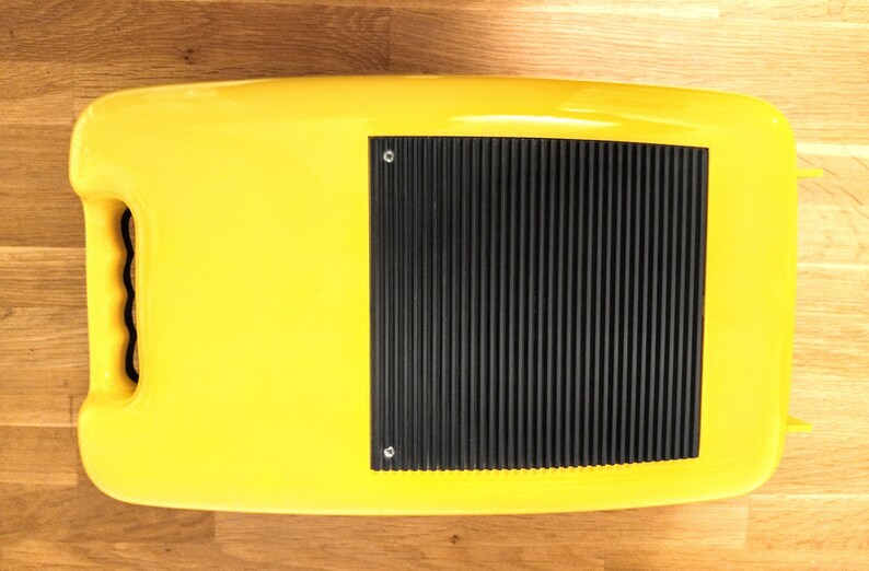 May include: A yellow plastic case with a black rubberised panel on the top. The panel has a series of raised lines and two small screws.