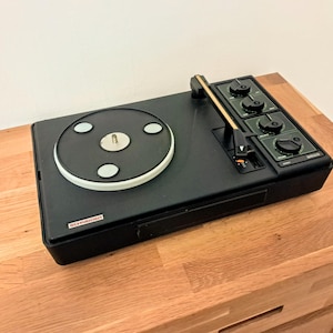 Puede incluir: Un tocadiscos negro con un plato blanco y un brazo de lectura dorado. El tocadiscos tiene una serie de botones y perillas en el lateral. El tocadiscos está sobre una superficie de madera.